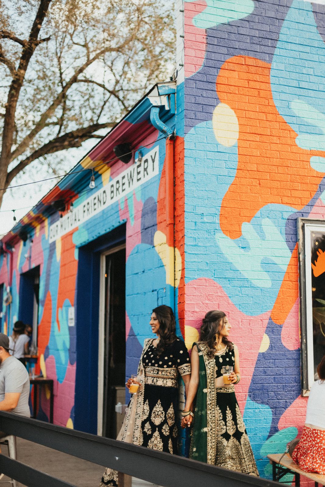 Two women in embroidered formal outfits holding hands in front of a brightly painted brewery mural