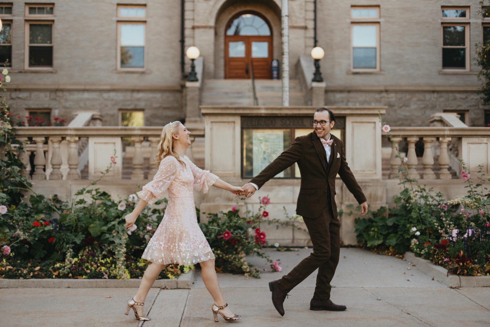 Engaged couple holding hands and walking past blooming flower beds in front of a historic building