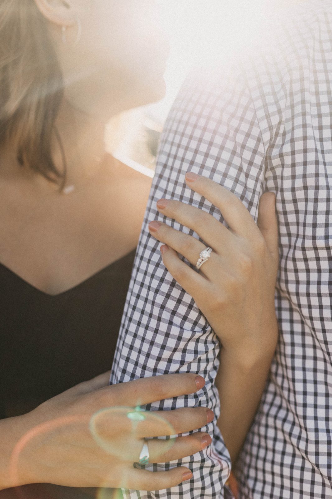Close-up of an engagement ring on a hand resting on her fiancé's checkered shirt in warm sunlight