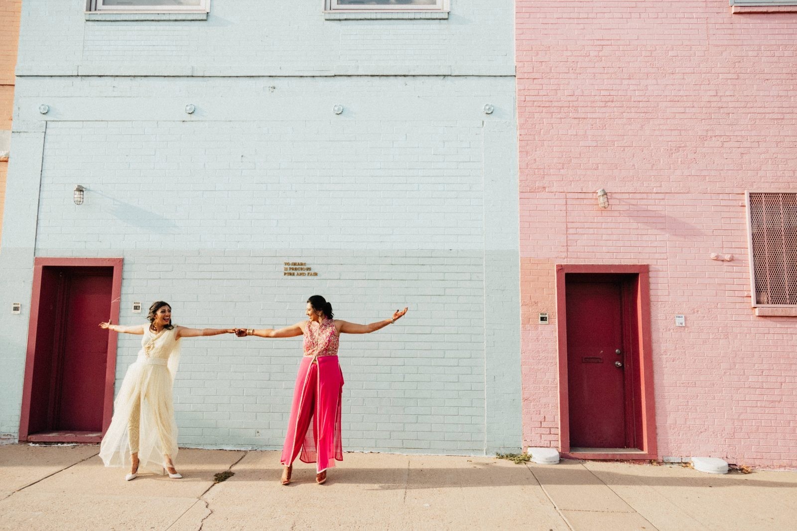 Two women in bright outfits reaching toward each other in front of pastel painted brick walls