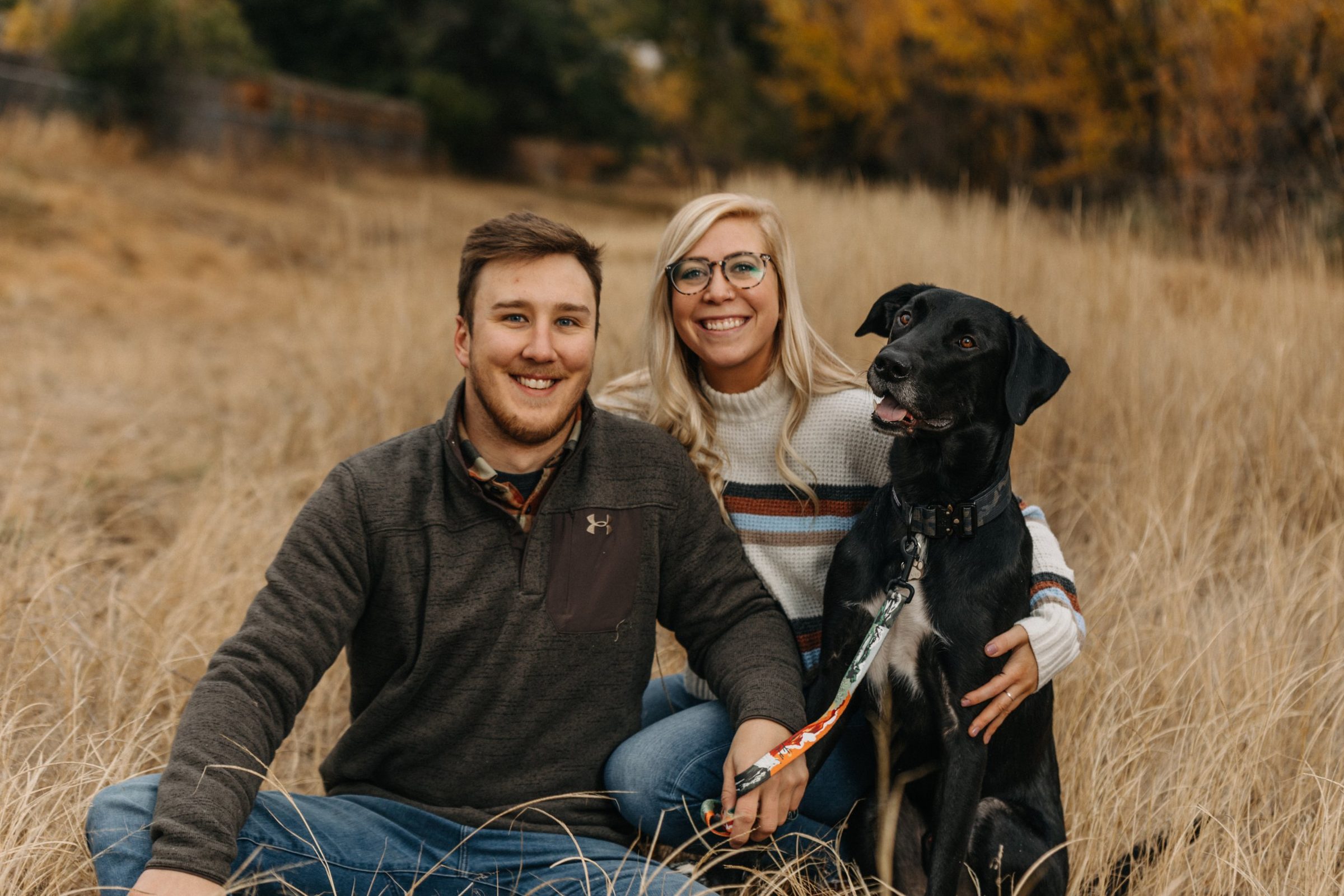 Smiling couple with their black dog sitting in tall golden grass among fall foliage