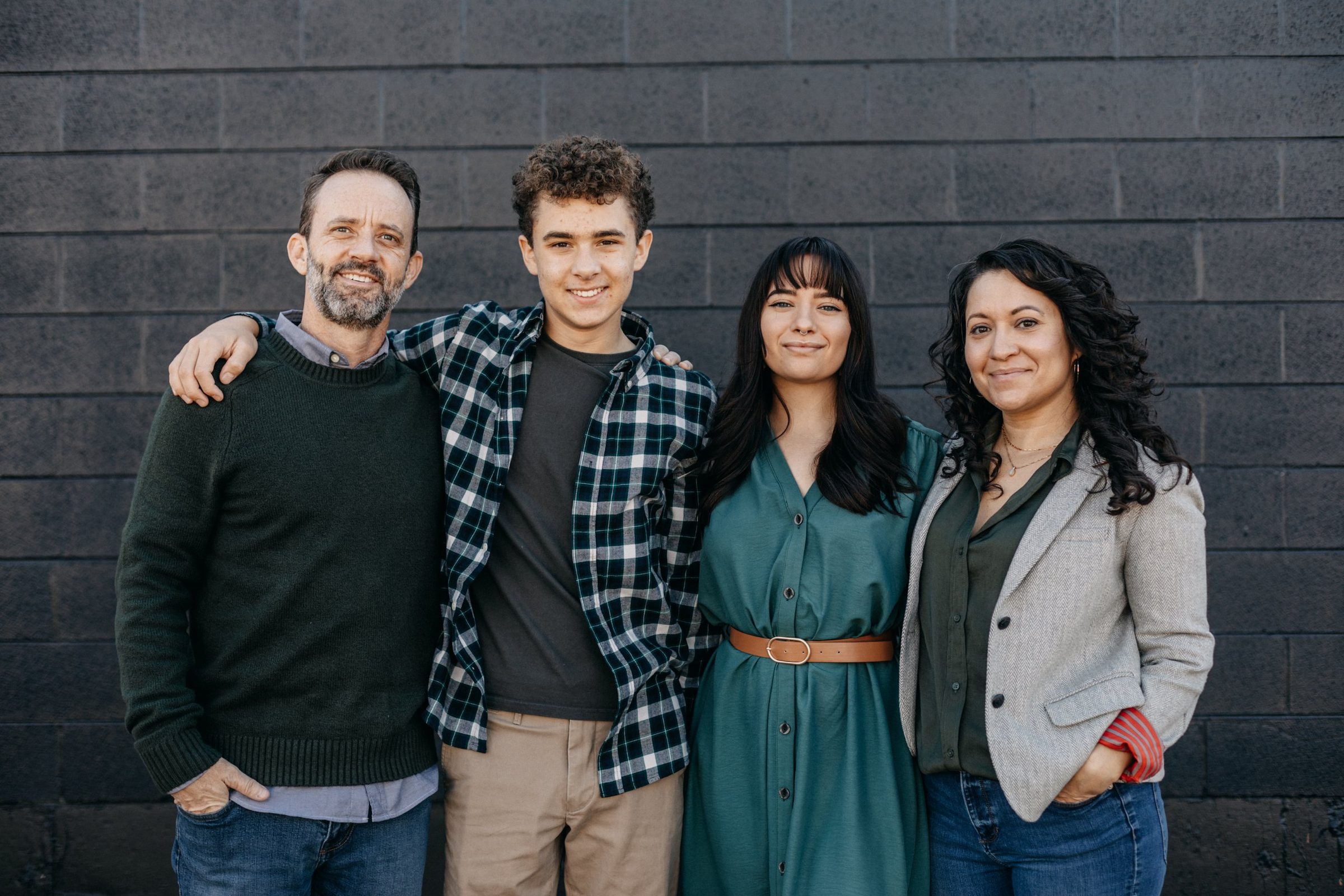 Family of four standing arm in arm in front of a dark painted brick wall