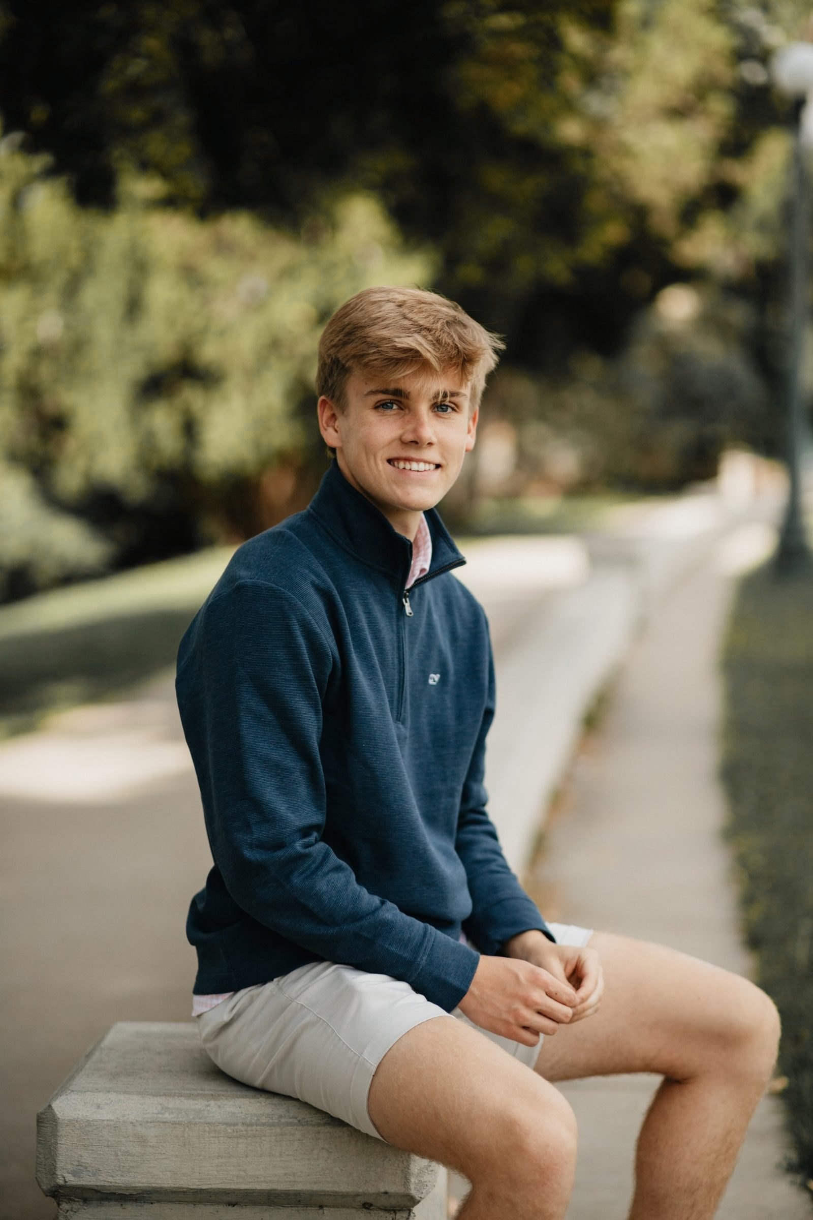 Smiling high school senior in a navy quarter-zip sitting on a stone pillar along a tree-lined path
