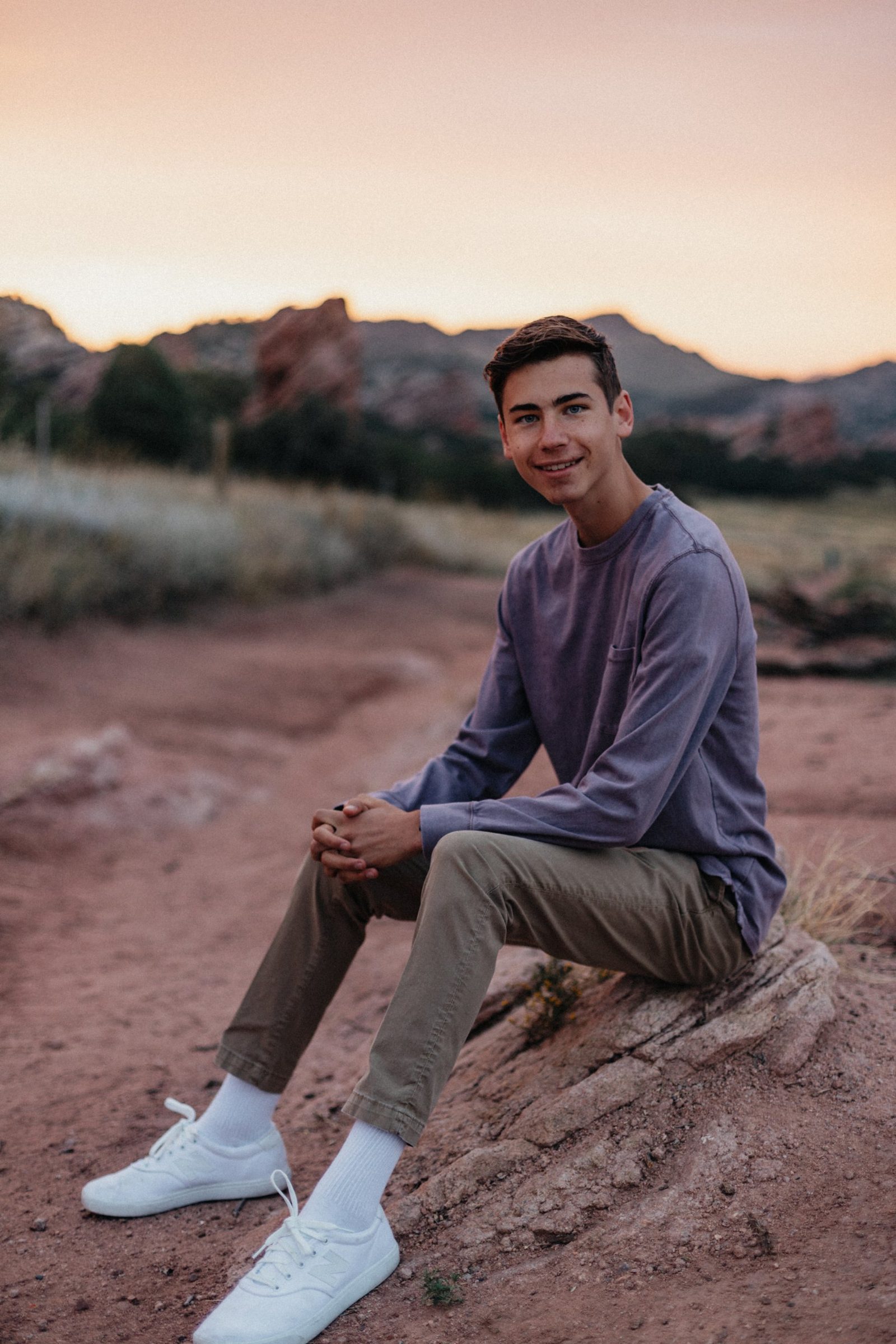 Senior in a purple long-sleeve shirt and white sneakers sitting on a rock at sunset with red rock formations behind