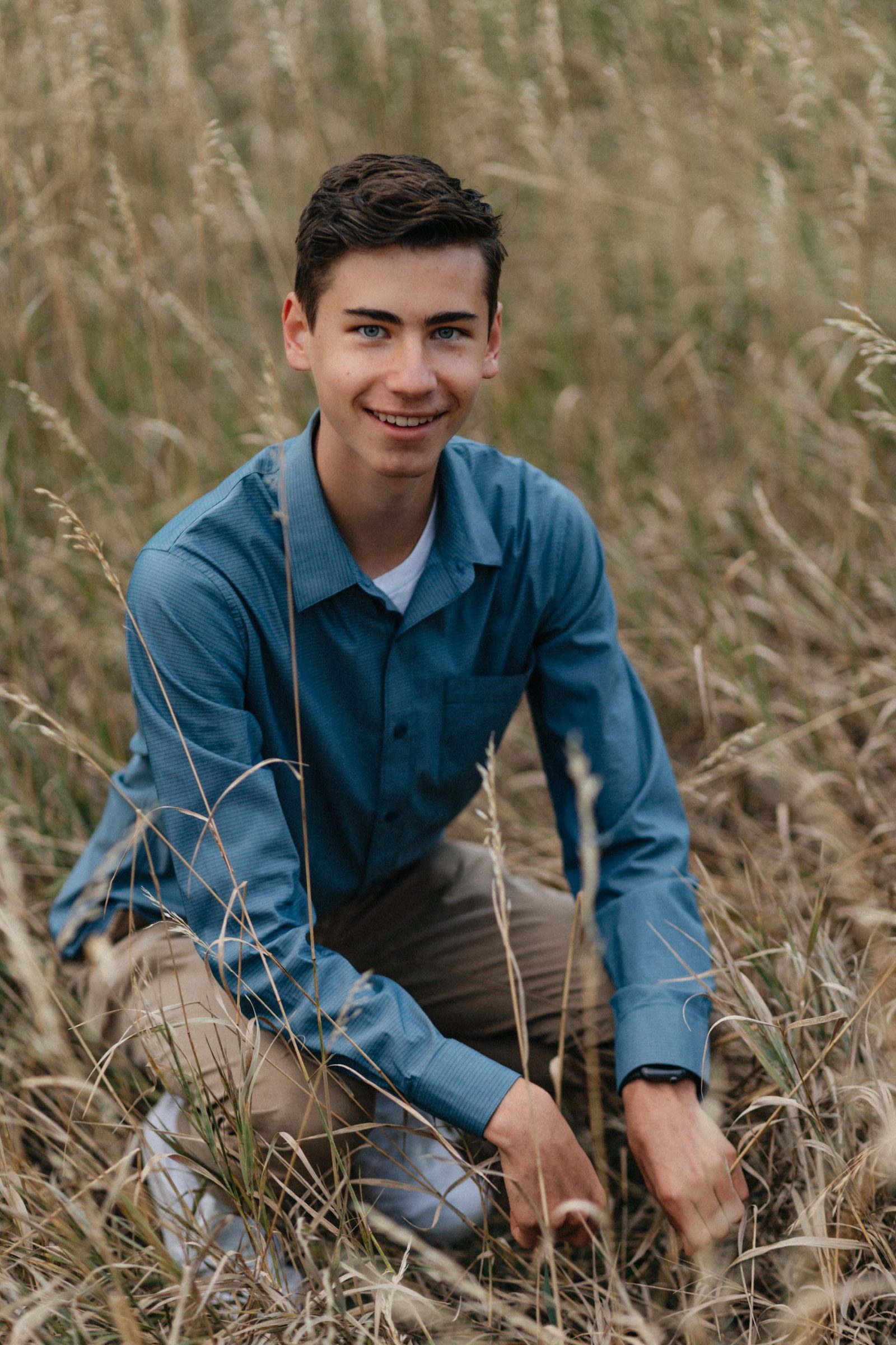 Smiling senior in a blue button-down crouching in tall golden grass