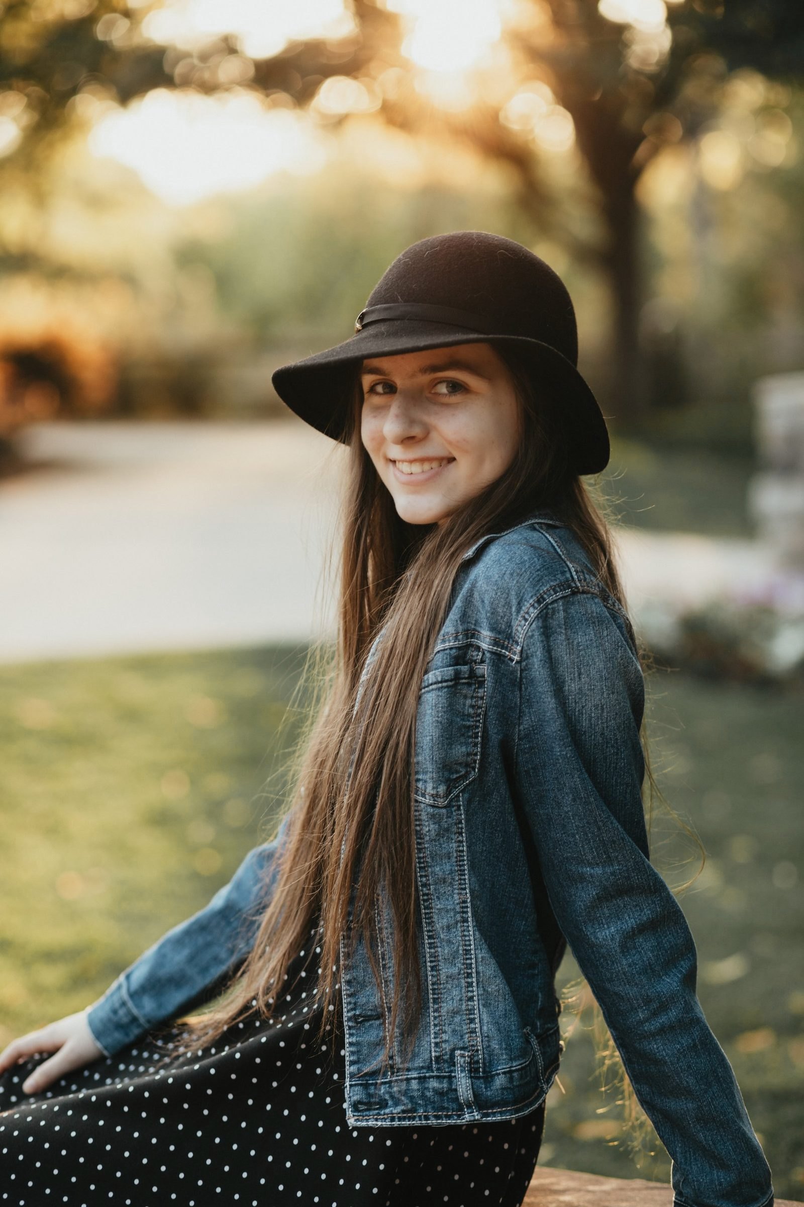 Senior girl in a black felt hat and denim jacket smiling over her shoulder in warm golden-hour light
