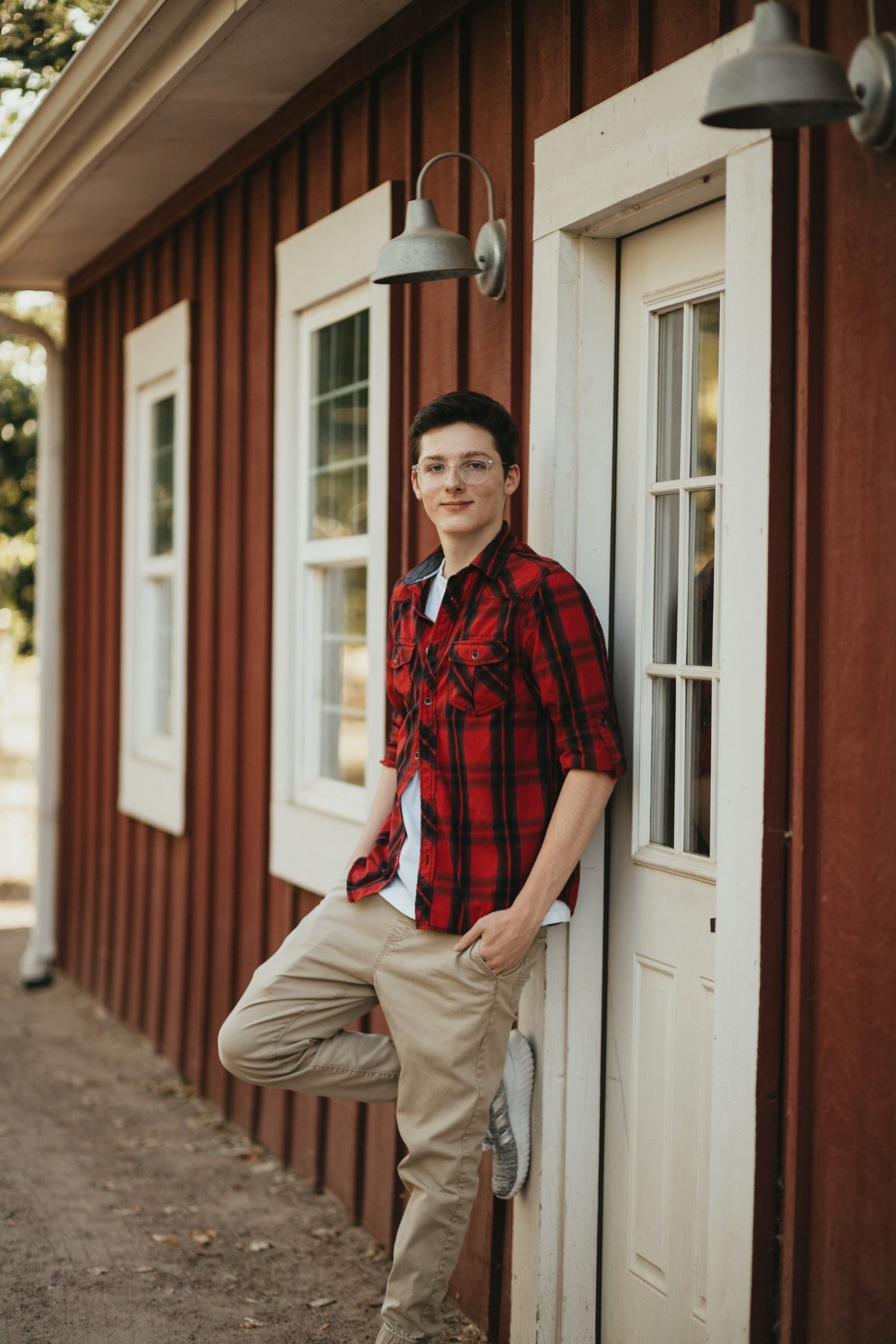Senior in clear glasses and a red plaid shirt leaning against a red barn wall beside a white door