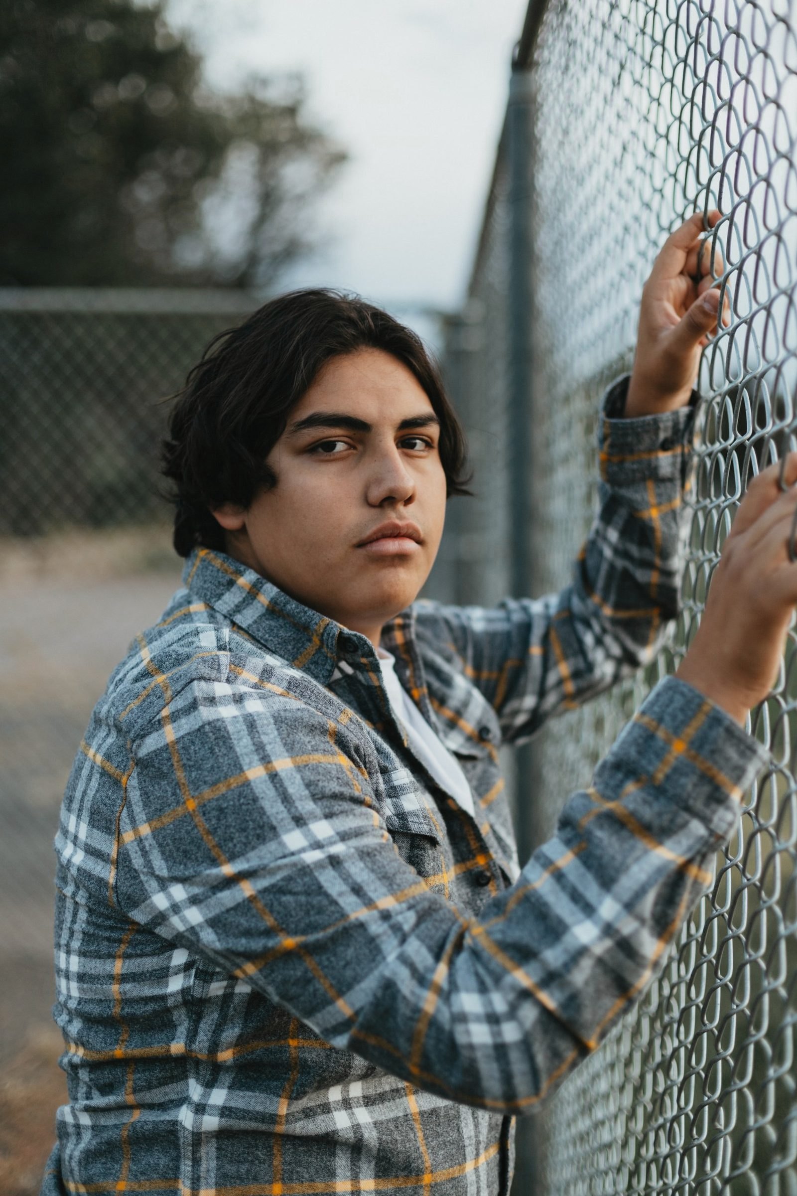 Senior in a gray and gold plaid flannel looking at the camera while gripping a chain-link fence