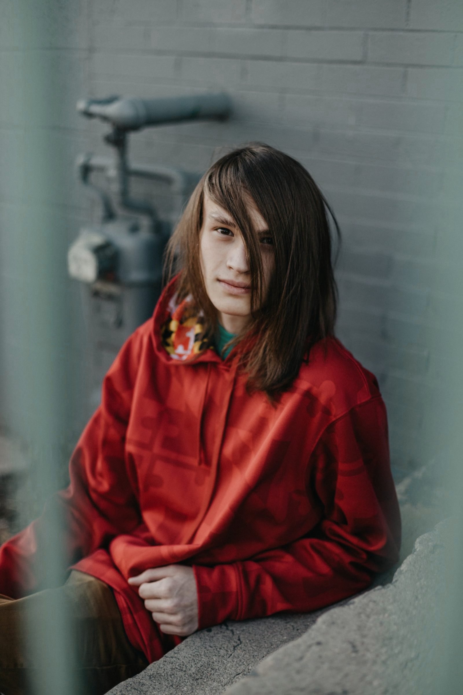 Senior with long brown hair in a red hoodie sitting against a gray brick wall