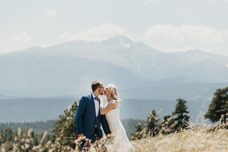 Bride and groom nearly kiss in a grassy mountain meadow with a hazy Rocky Mountain peak rising behind them