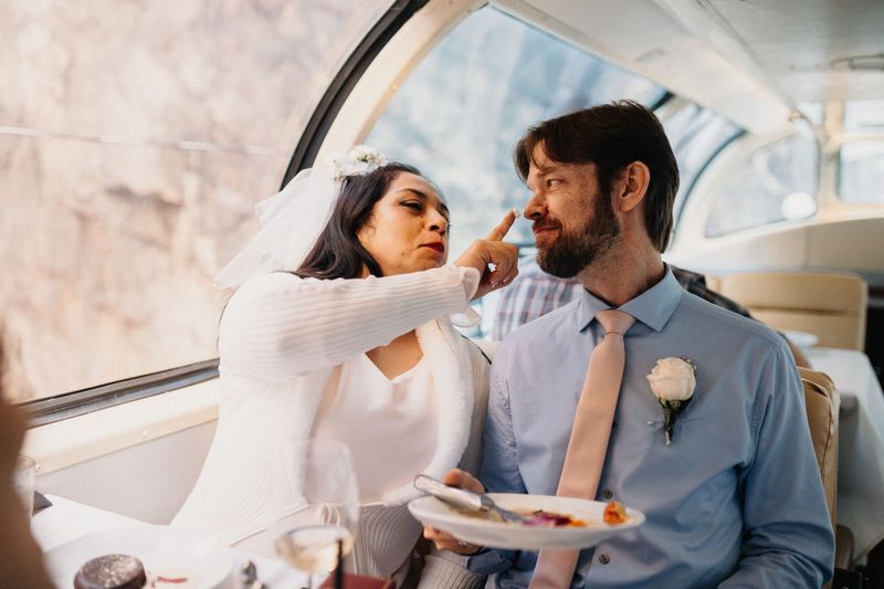 Bride playfully touches her husband's nose while they share dessert in a glass-roofed train car