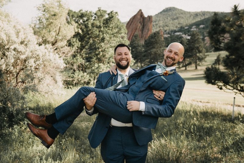 Groom laughing as he is carried in his friend's arms on a grassy hillside with red rocks behind