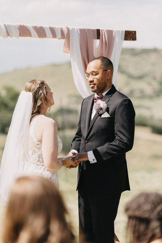 Bride and groom hold hands under a draped wooden arch during an outdoor hilltop ceremony