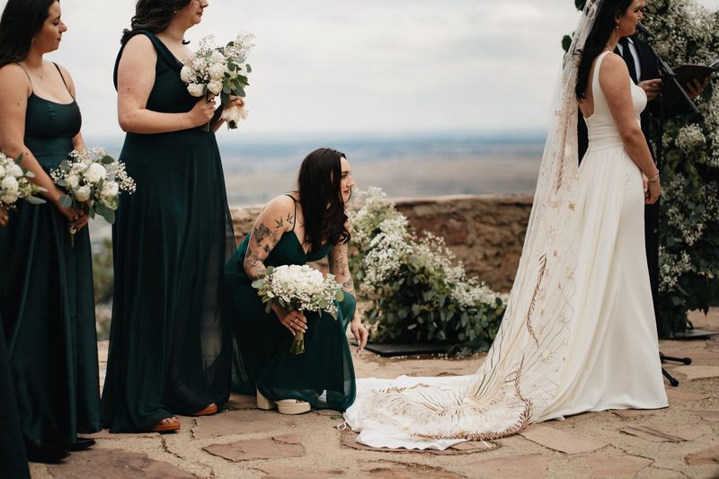 Bridesmaids in deep green dresses gather as the bride's long veil trails across a stone overlook