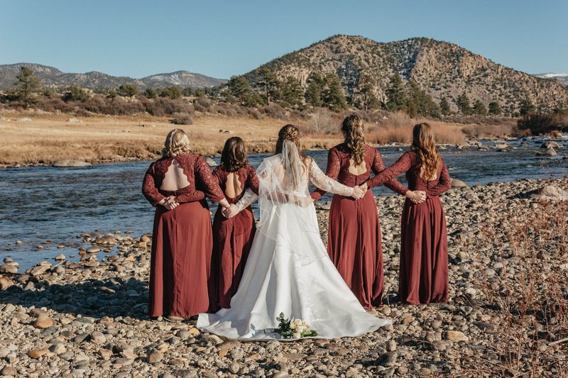Bride and four bridesmaids in burgundy dresses link arms facing a river and mountains