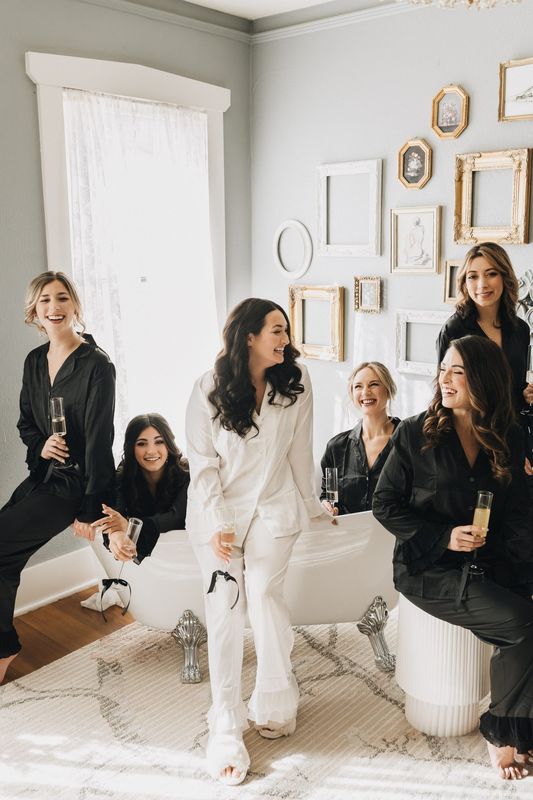 Bride in white pajamas laughs with her bridesmaids in black robes while getting ready