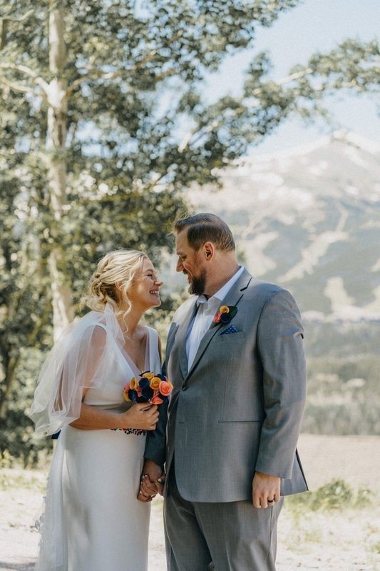 Smiling bride and groom hold hands with snowy ski slopes visible in the mountains behind them