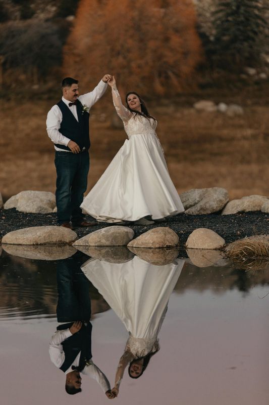 Groom twirls his bride on a rocky shoreline at dusk with a mirror-like reflection in the water