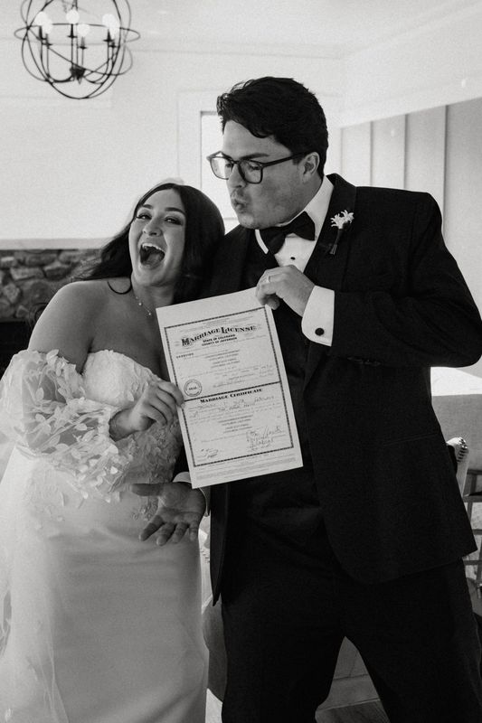 Black and white photo of a laughing bride and groom holding up their marriage license