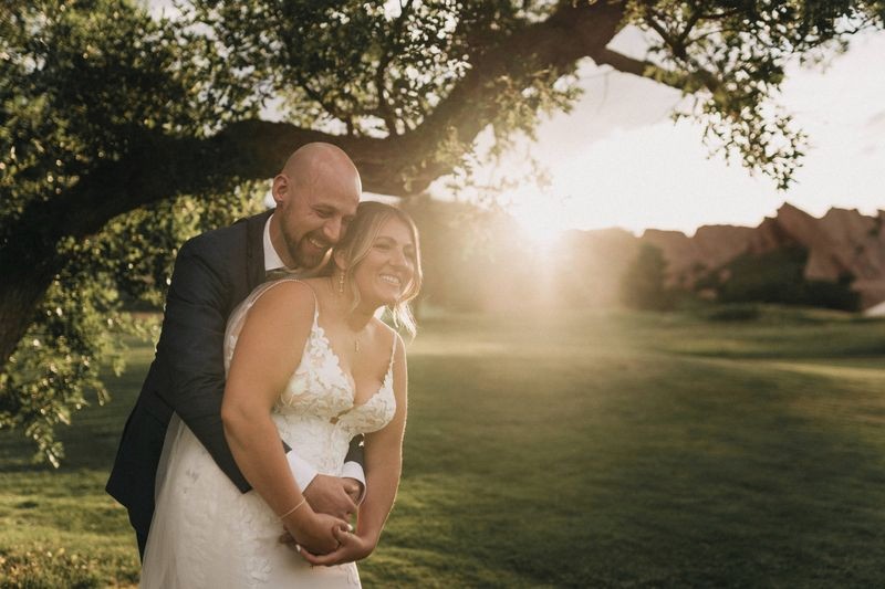 Groom hugs his bride from behind as the sun sets over red rocks on a golf course