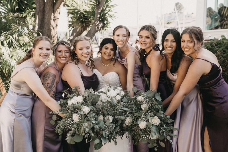 Bride and eight bridesmaids in lavender dresses laughing around a large bouquet