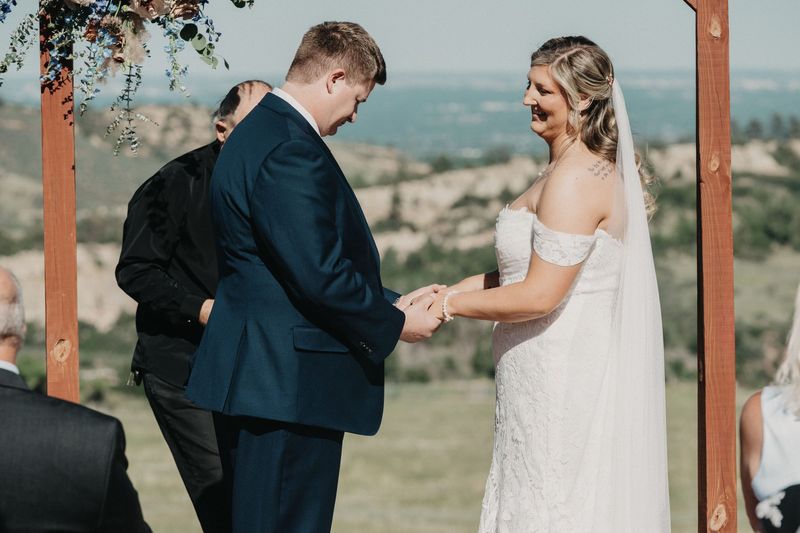 Bride and groom smile and hold hands under a rustic wooden arch above rolling hills