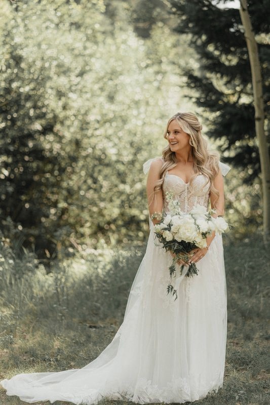 Bride holding a white bouquet and smiling in a sunny aspen grove