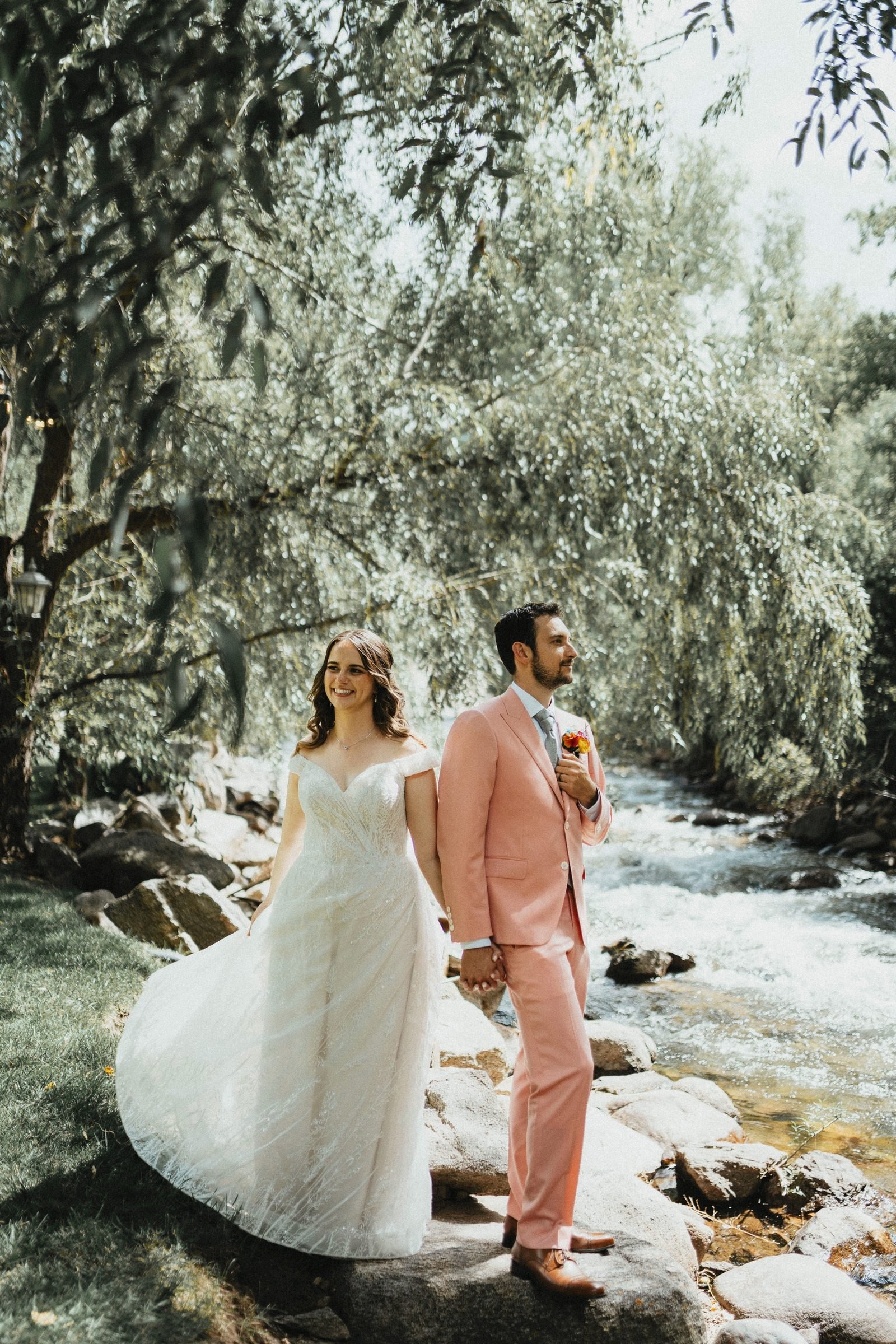Bride and groom in a peach suit hold hands beside a rushing mountain creek under willow trees