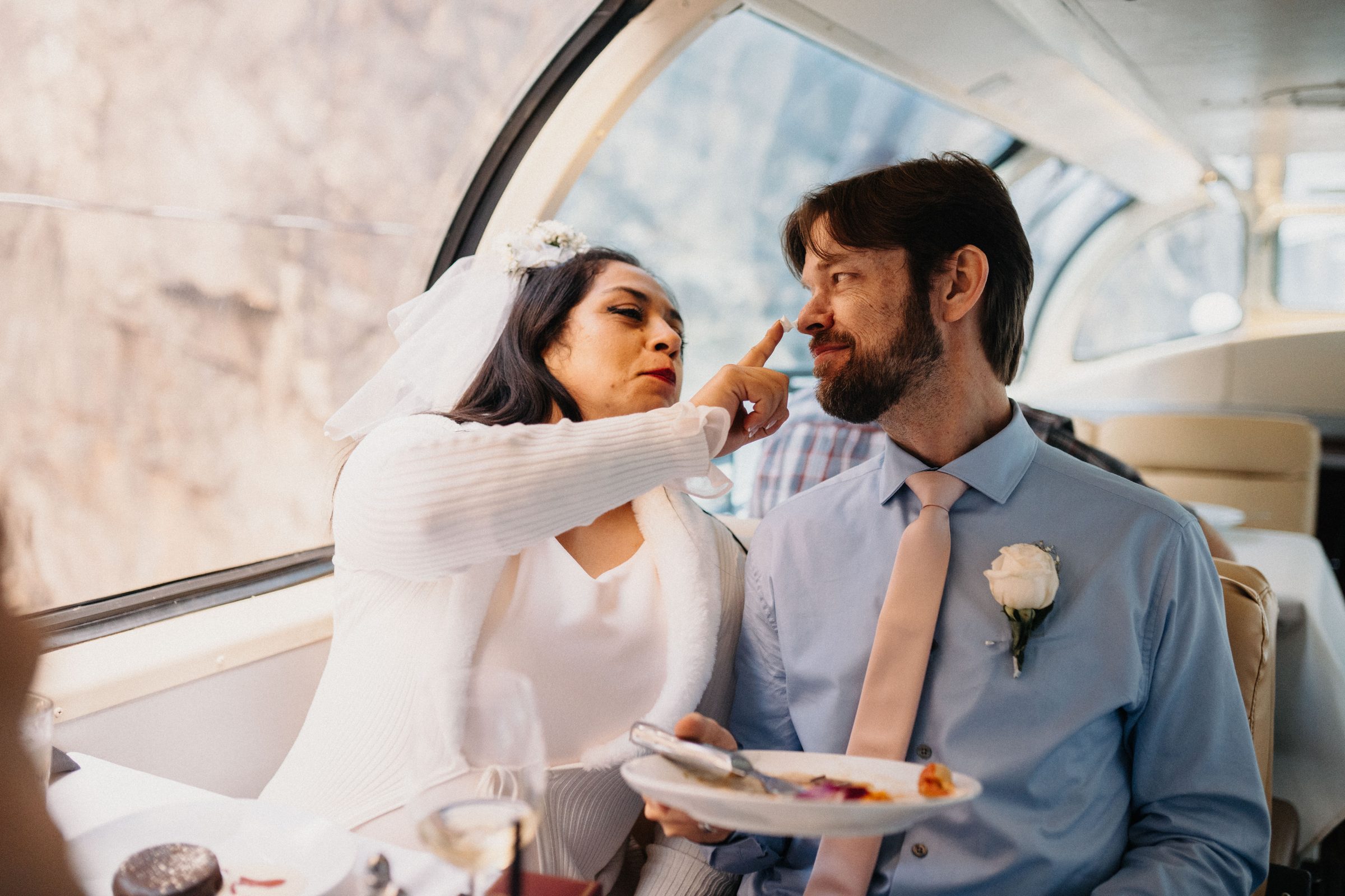 Bride playfully touches her husband's nose while they share dessert in a glass-roofed train car