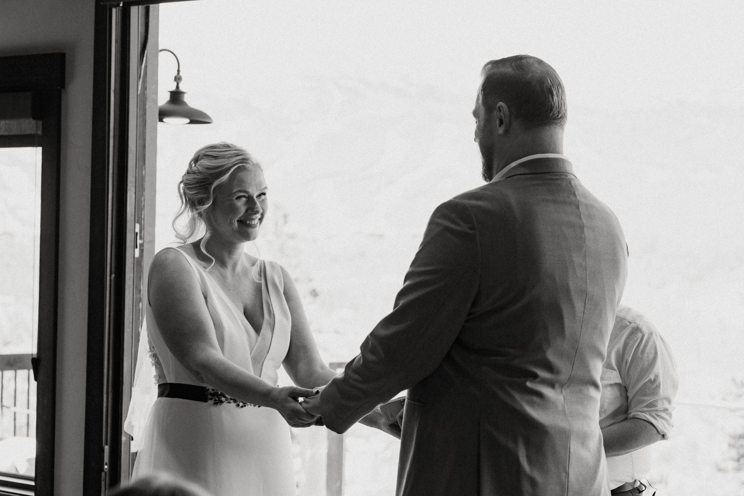 Black and white photo of a bride and groom holding hands during their ceremony by a bright window