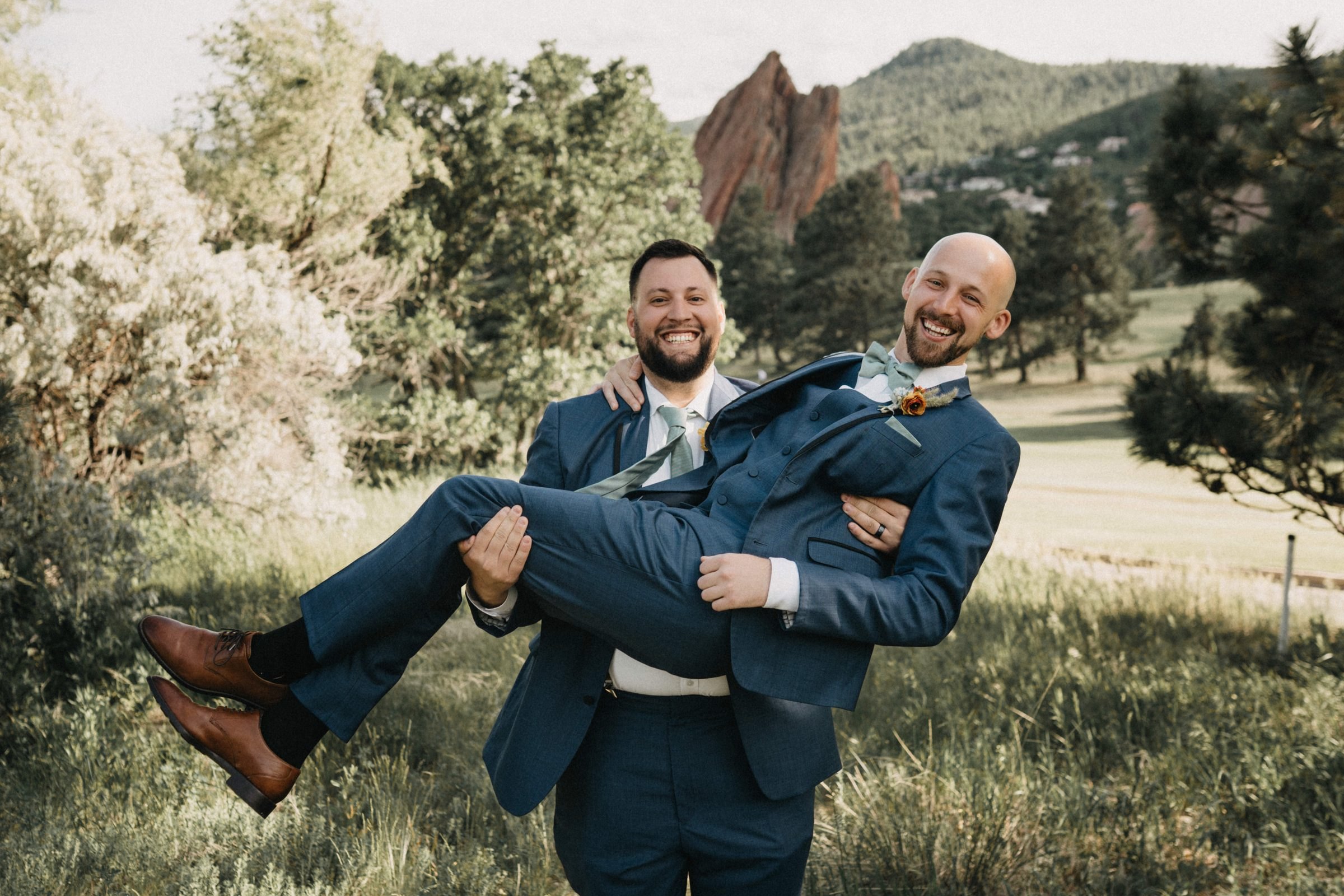 Groom laughing as he is carried in his friend's arms on a grassy hillside with red rocks behind