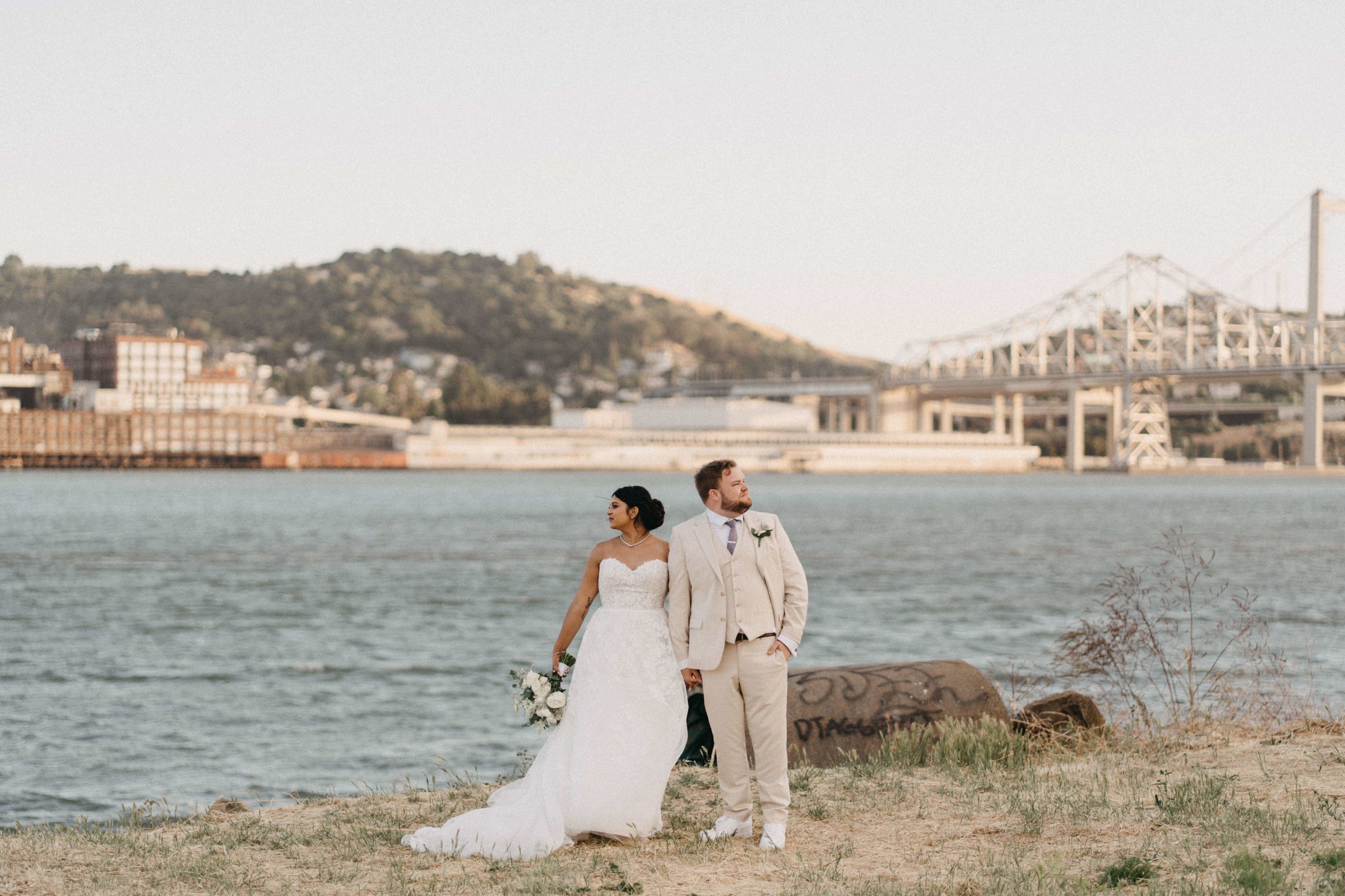 Bride and groom standing together on a rocky shoreline at sunset with a bridge in the distance