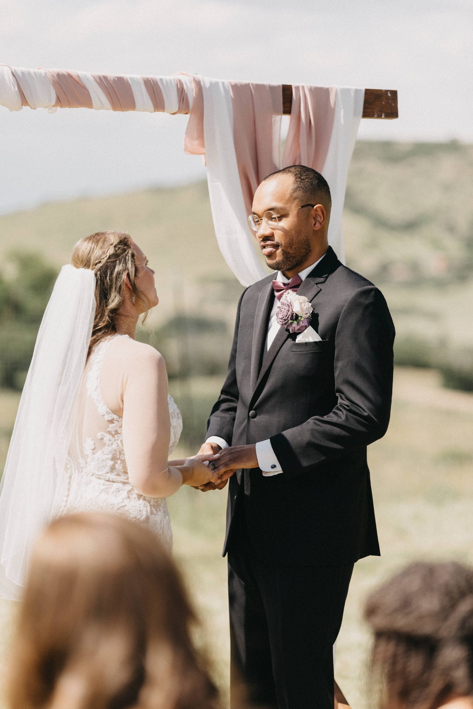 Bride and groom hold hands under a draped wooden arch during an outdoor hilltop ceremony