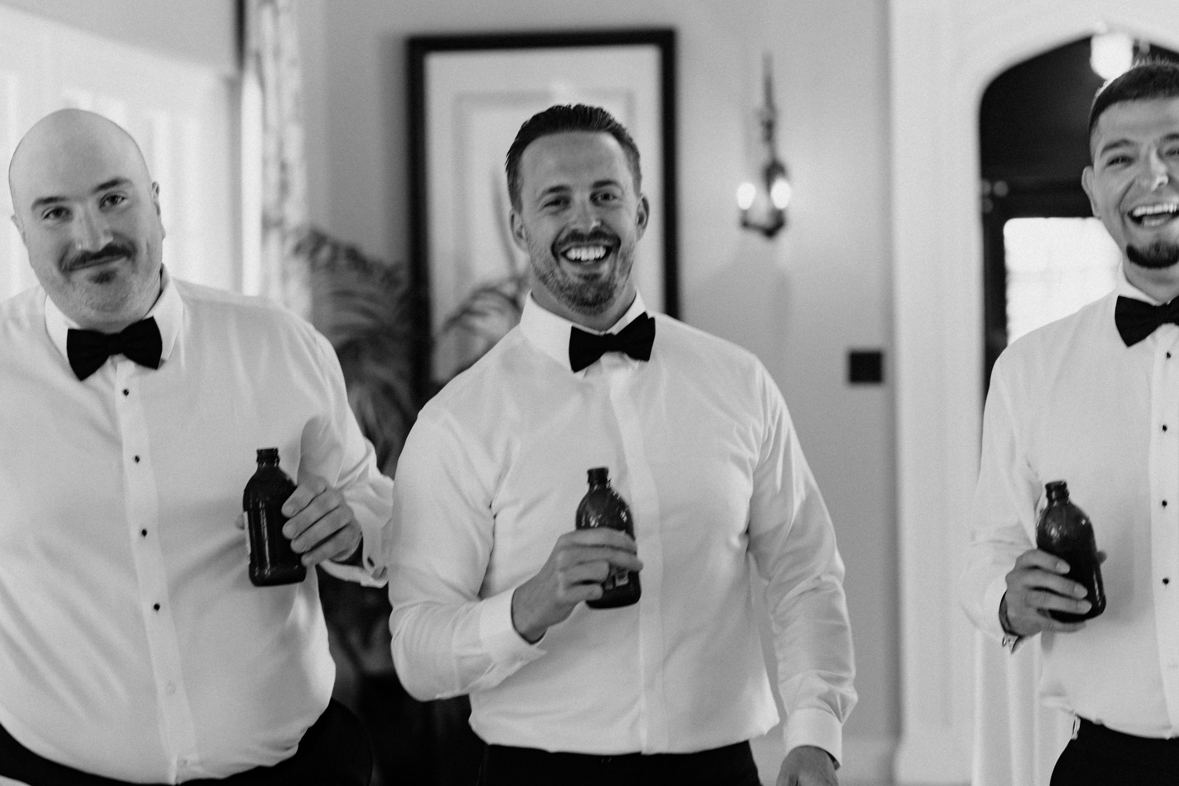 Black and white photo of groomsmen in bow ties laughing and holding drinks before the ceremony