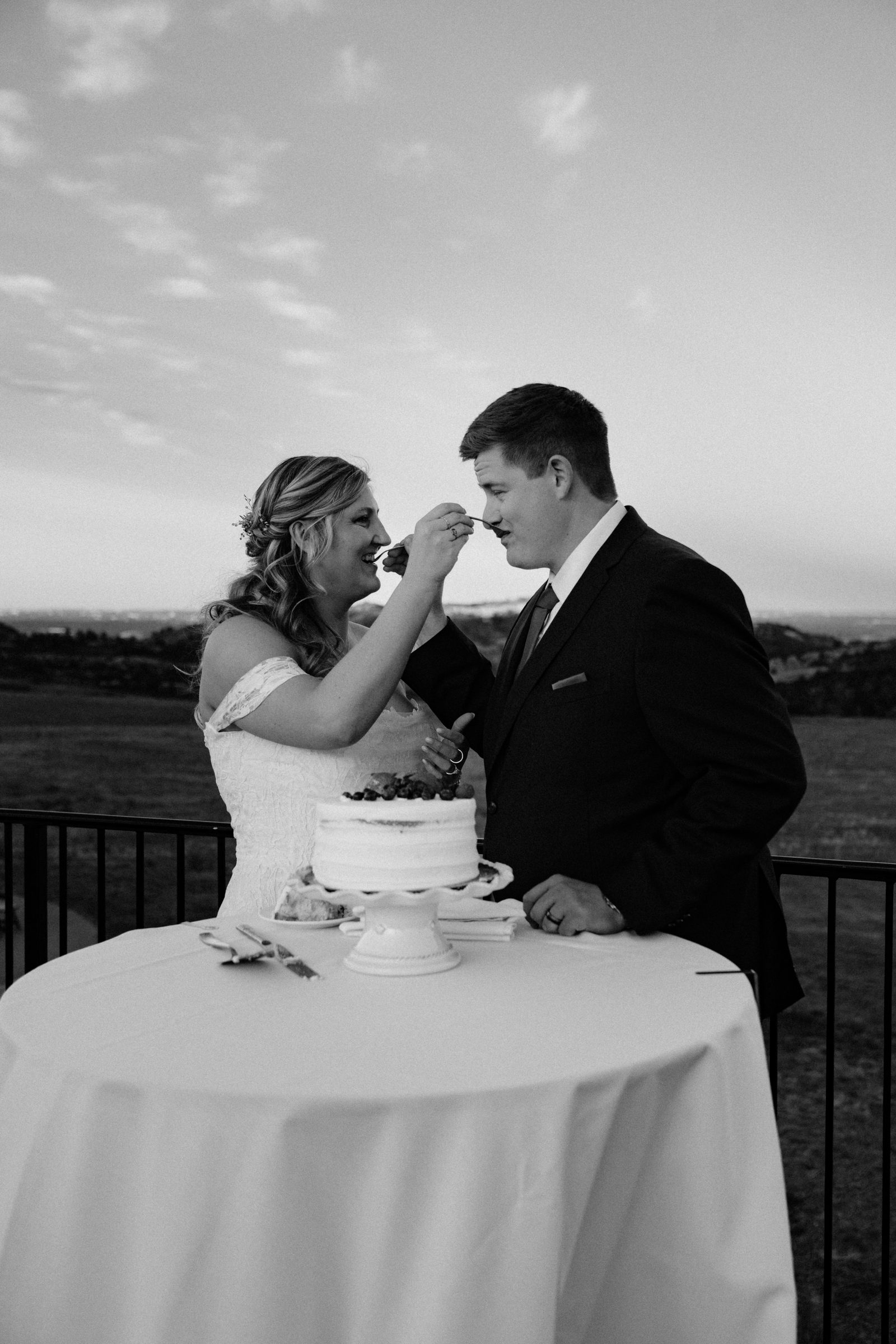 Black and white photo of a bride feeding her groom a bite of wedding cake at a table overlooking the plains