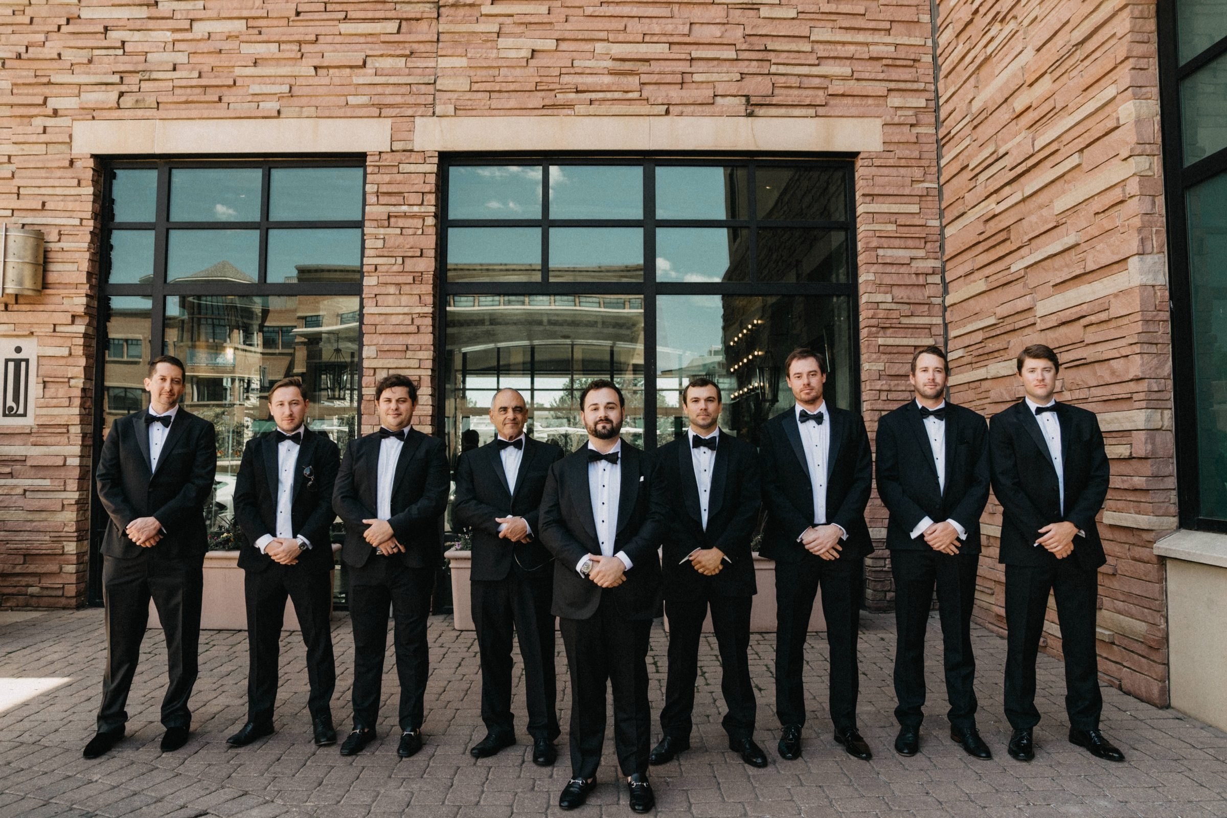 Nine groomsmen in black tuxedos standing in a line in front of a brick and glass building