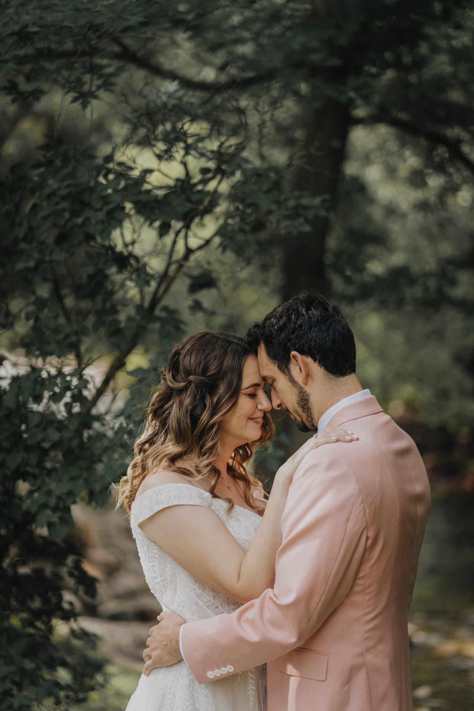 Bride and groom touch foreheads in a quiet moment among green trees