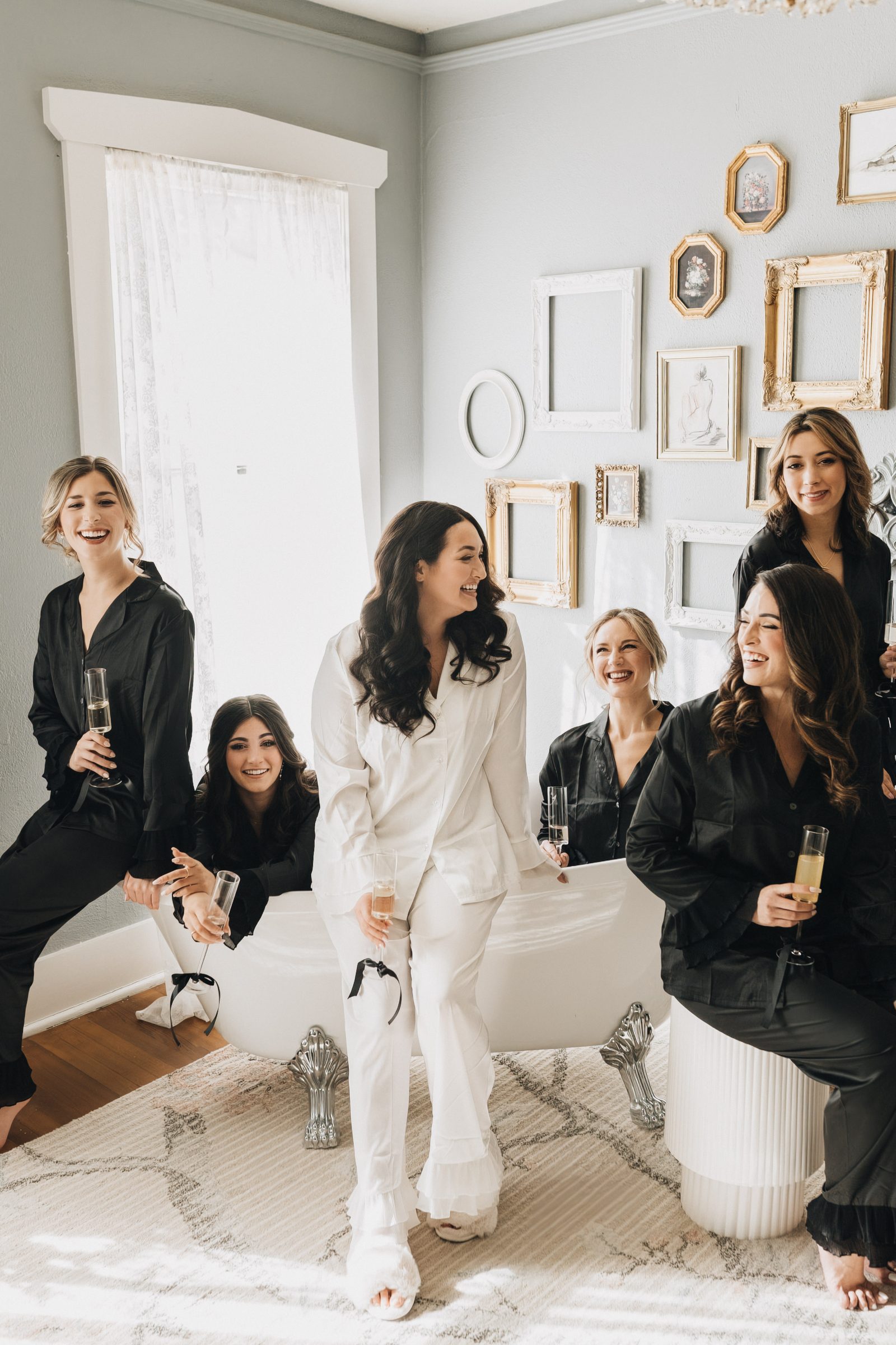 Bride in white pajamas laughs with her bridesmaids in black robes while getting ready