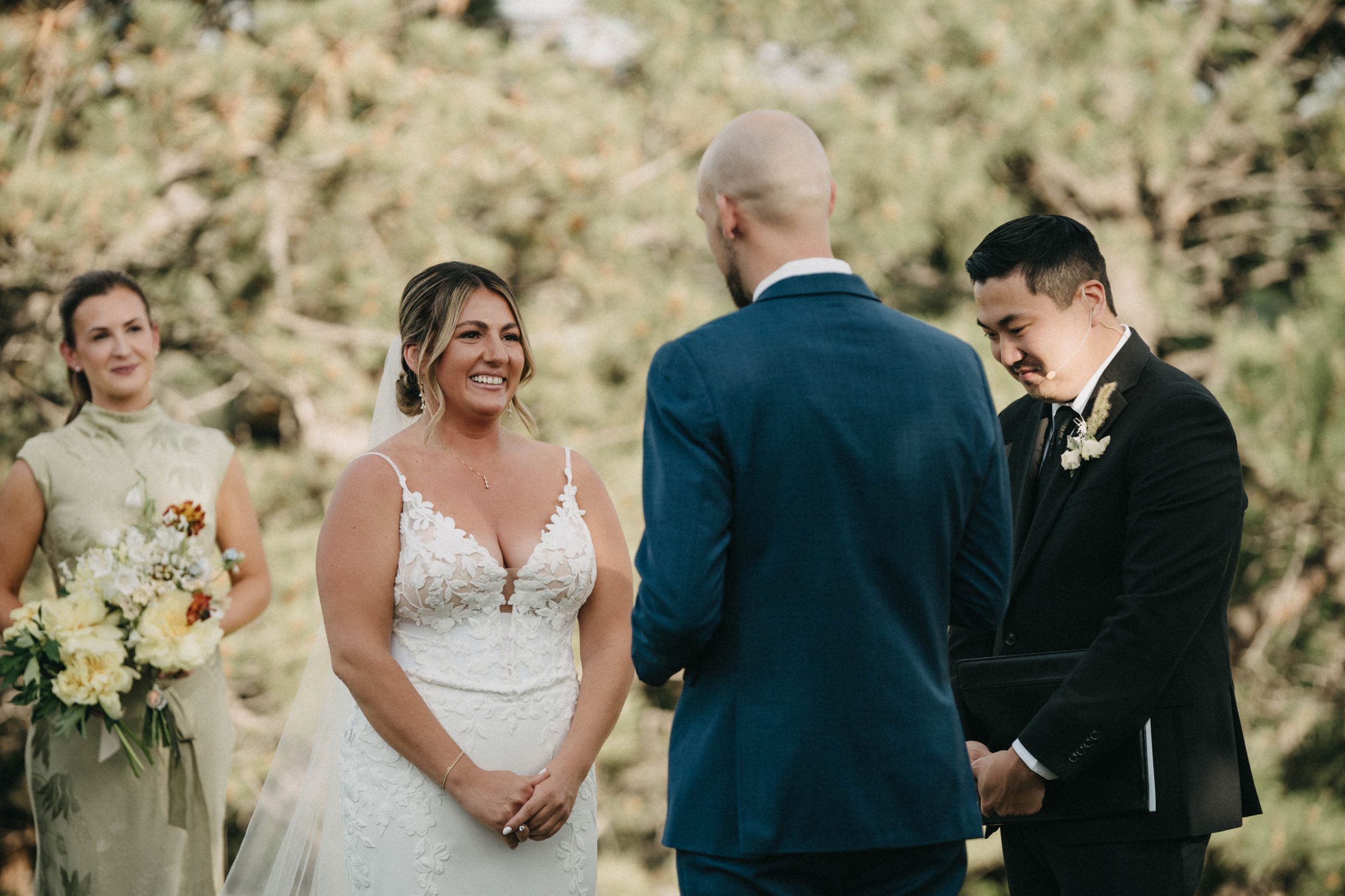 Bride smiles as her groom and officiant stand together during an outdoor pine-forest ceremony