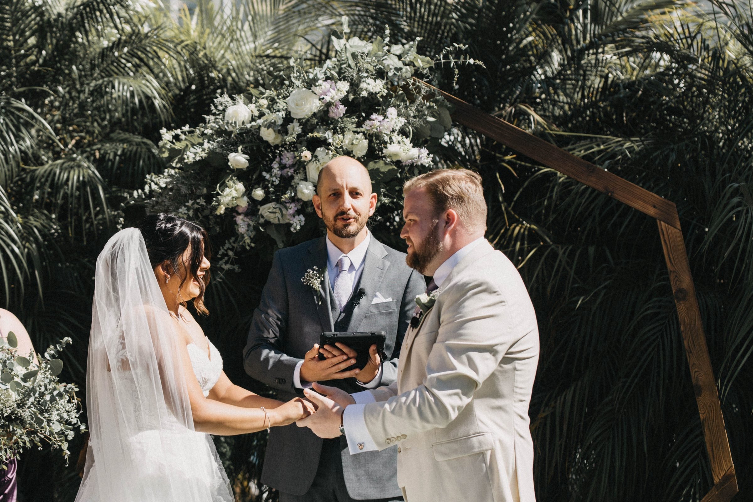 Bride and groom hold hands under a floral arch with palm trees as the officiant reads from a tablet