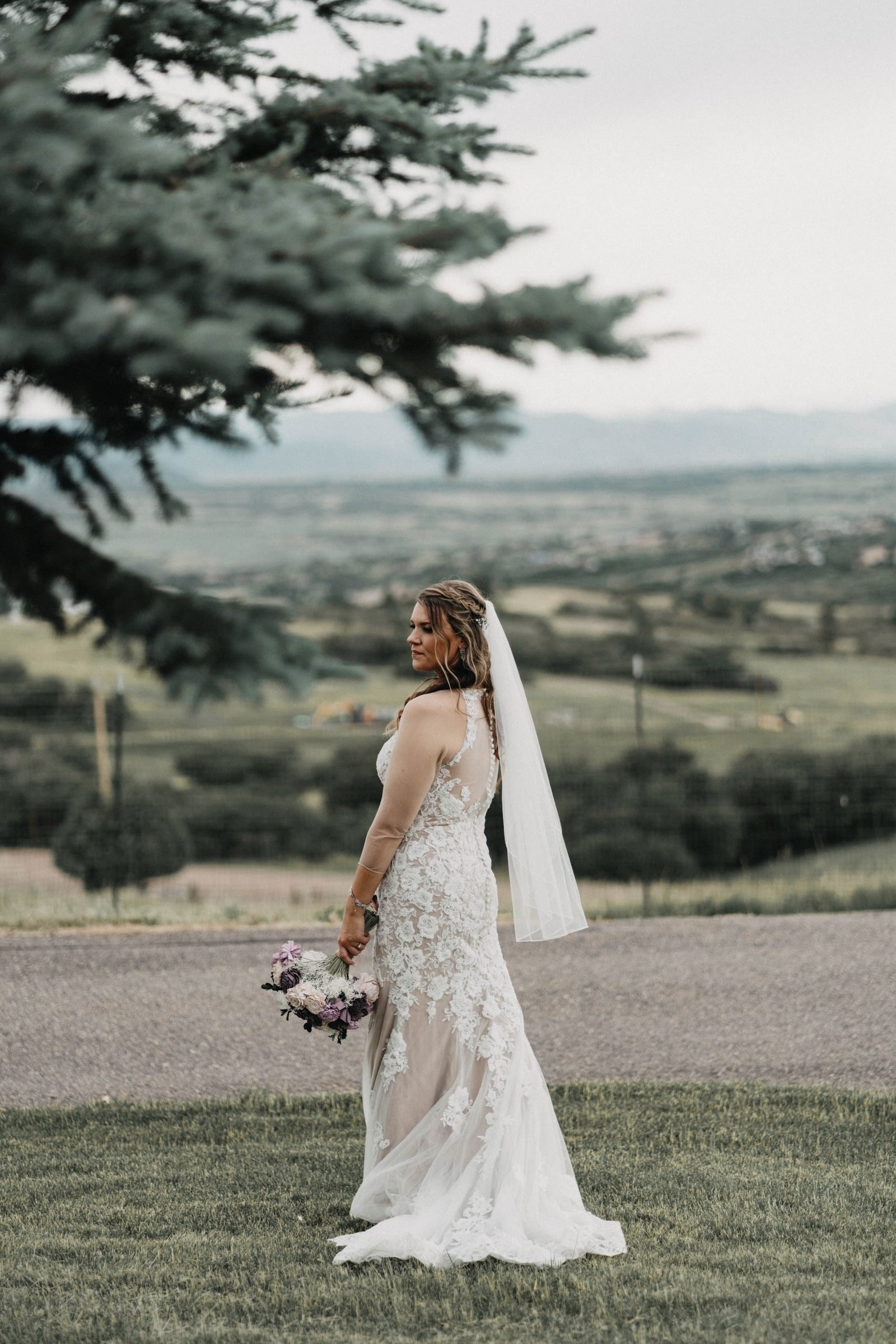 Bride in a lace gown holding a bouquet, looking out over green foothills and farmland