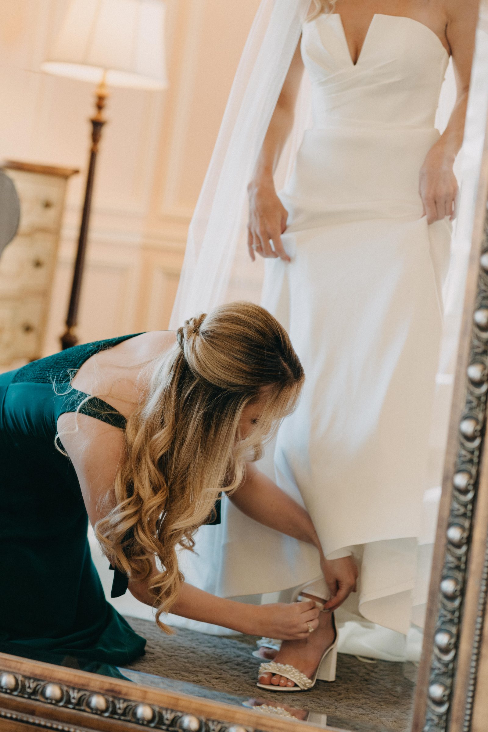 Bridesmaid helping the bride into her shoes in front of a gold-framed mirror