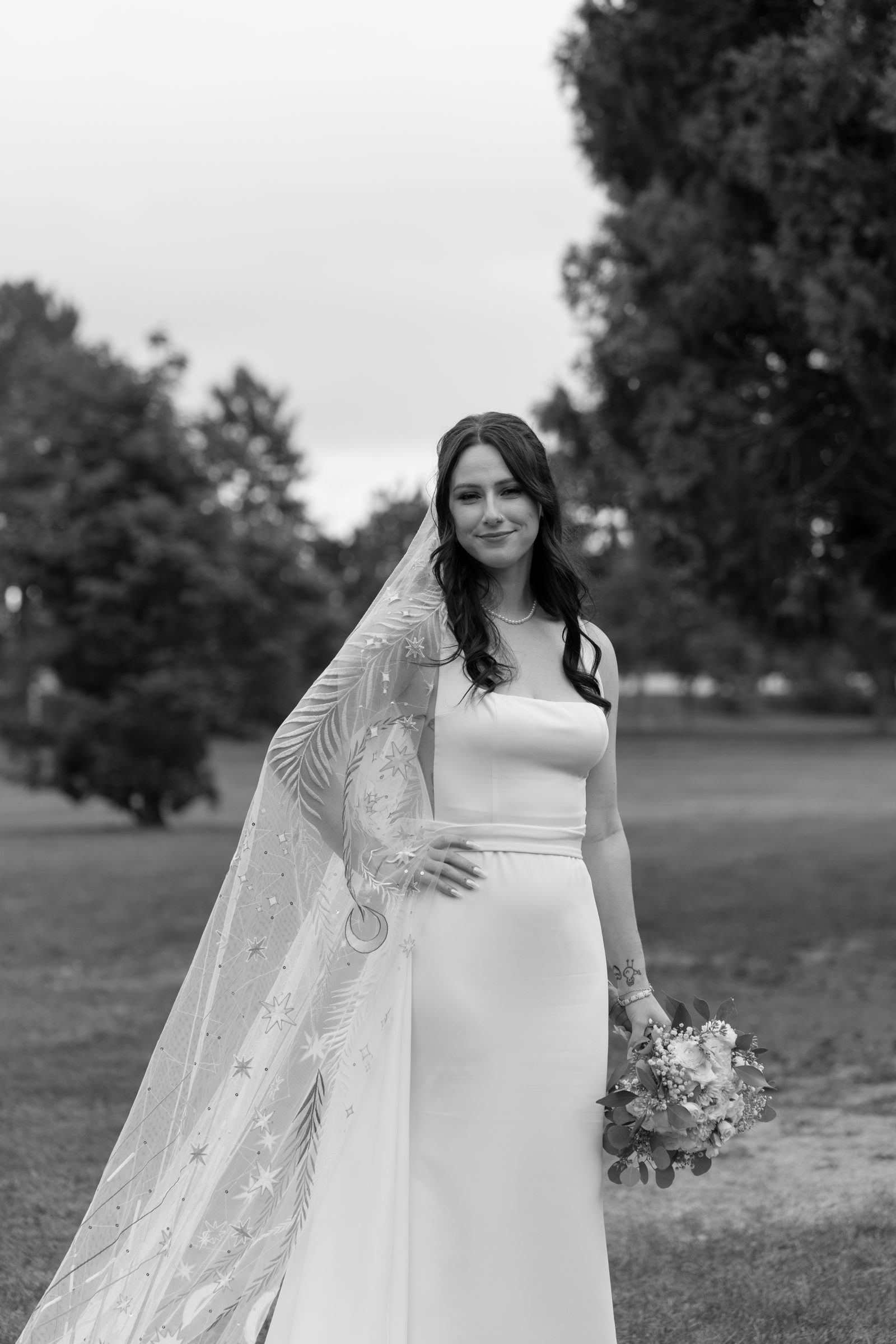 Black and white portrait of a bride in a patterned lace veil holding a bouquet in a park