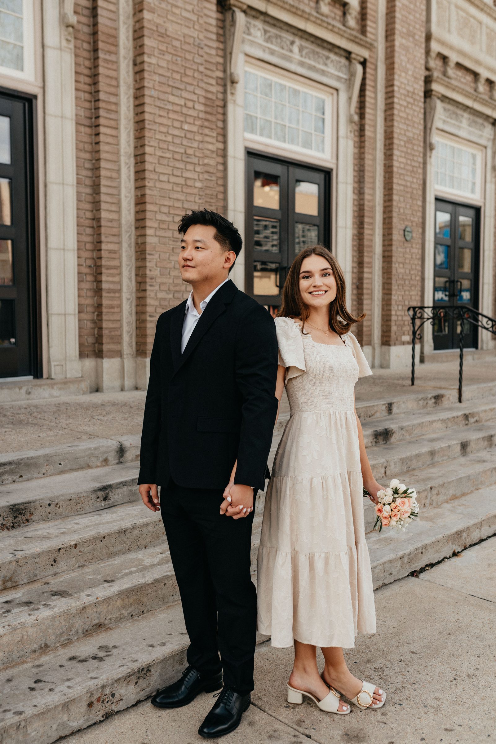 Couple holding hands on the steps of a brick building, the bride holding a small bouquet