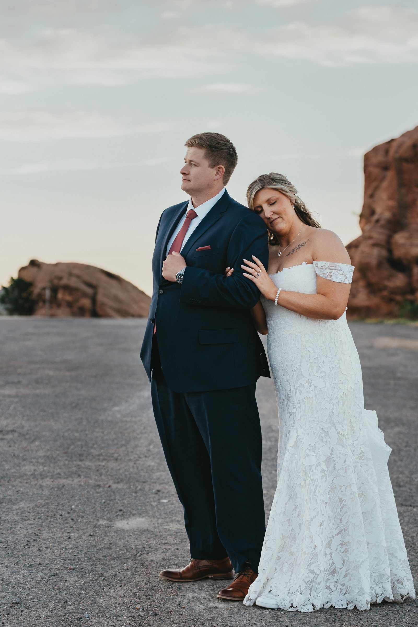 Groom and bride lean together on a gravel road with red sandstone rocks at sunset