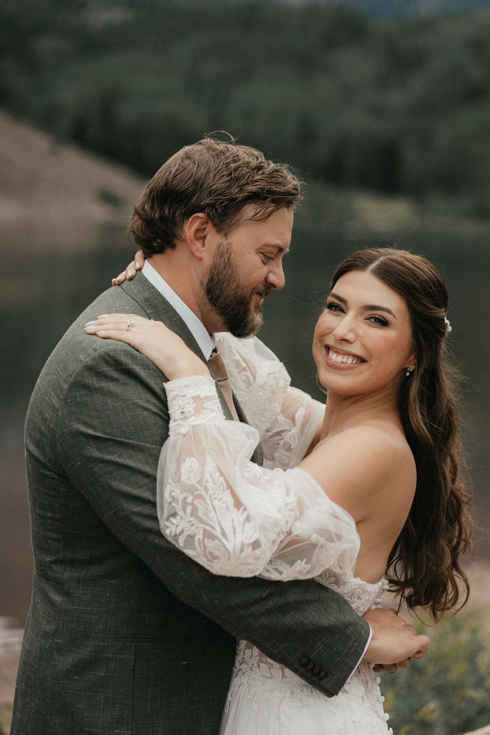 Bride and groom embrace and smile beside a mountain lake