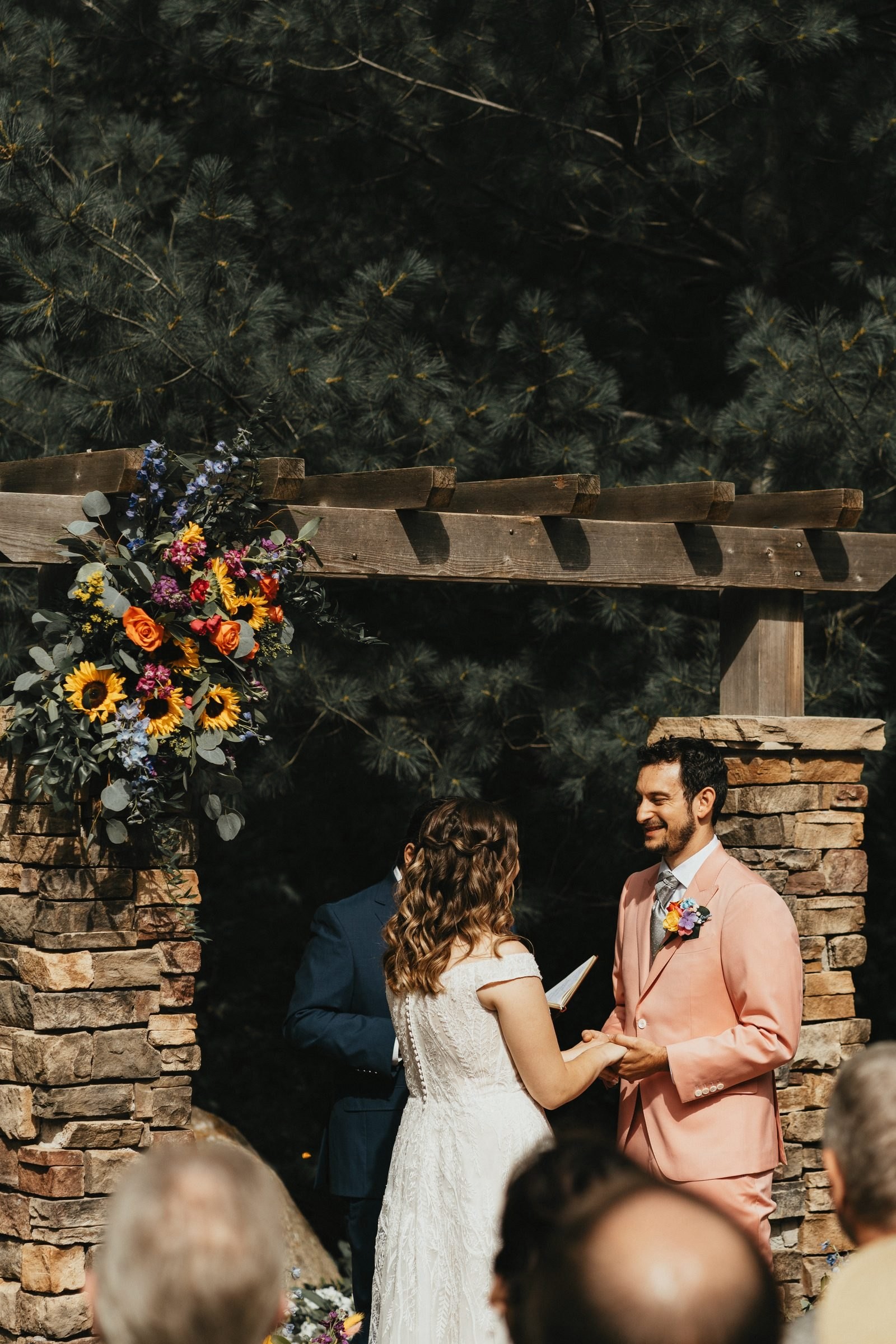 Bride and groom in a peach suit exchange vows under a stone and timber arch with sunflowers