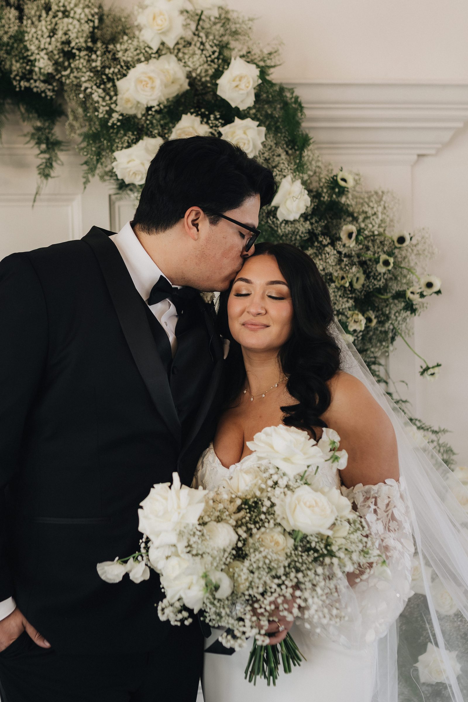 Groom kisses his bride on the forehead as she holds a white bouquet beneath a floral arch