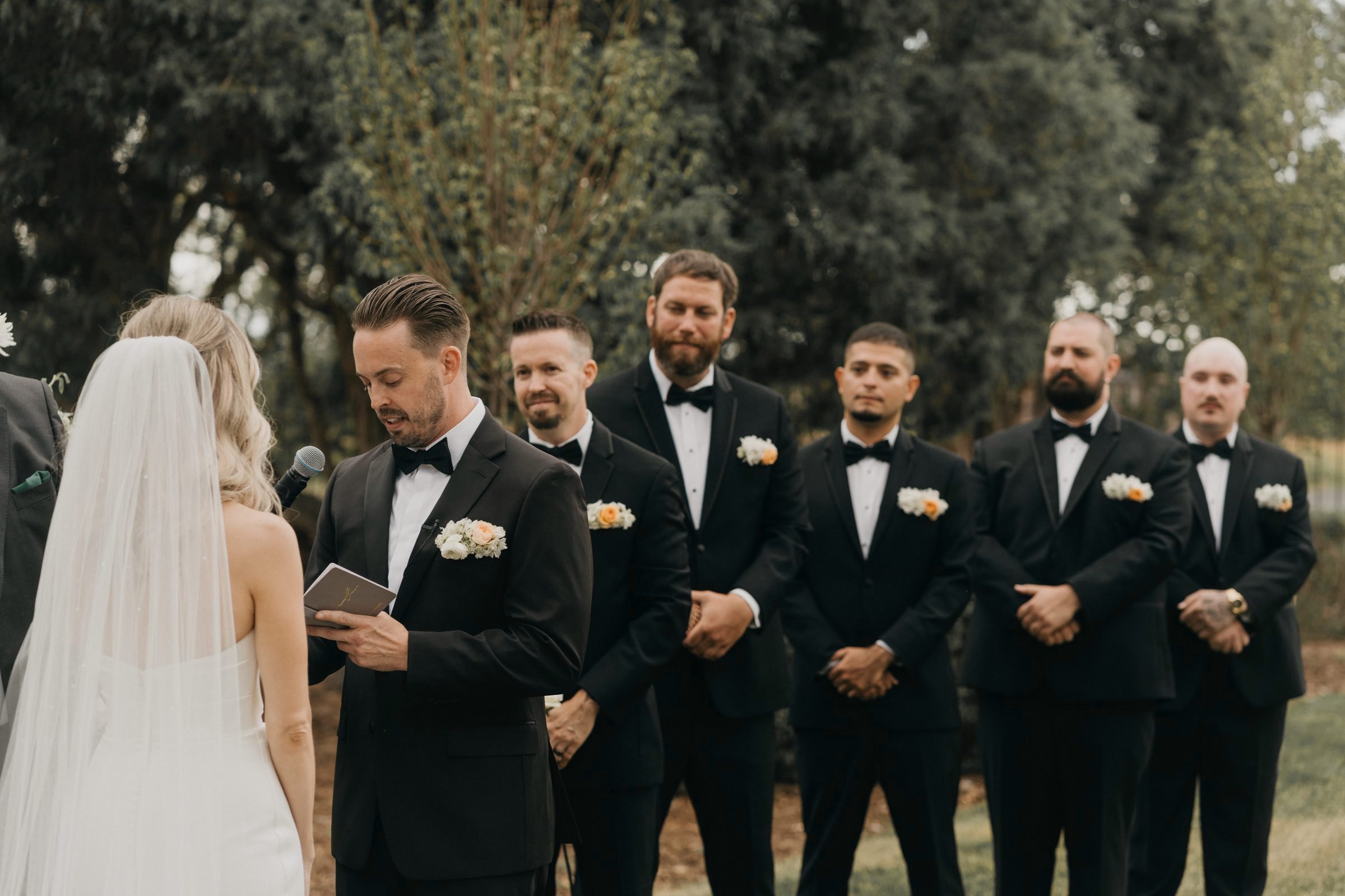 Groom reads his vows in front of six groomsmen during an outdoor ceremony