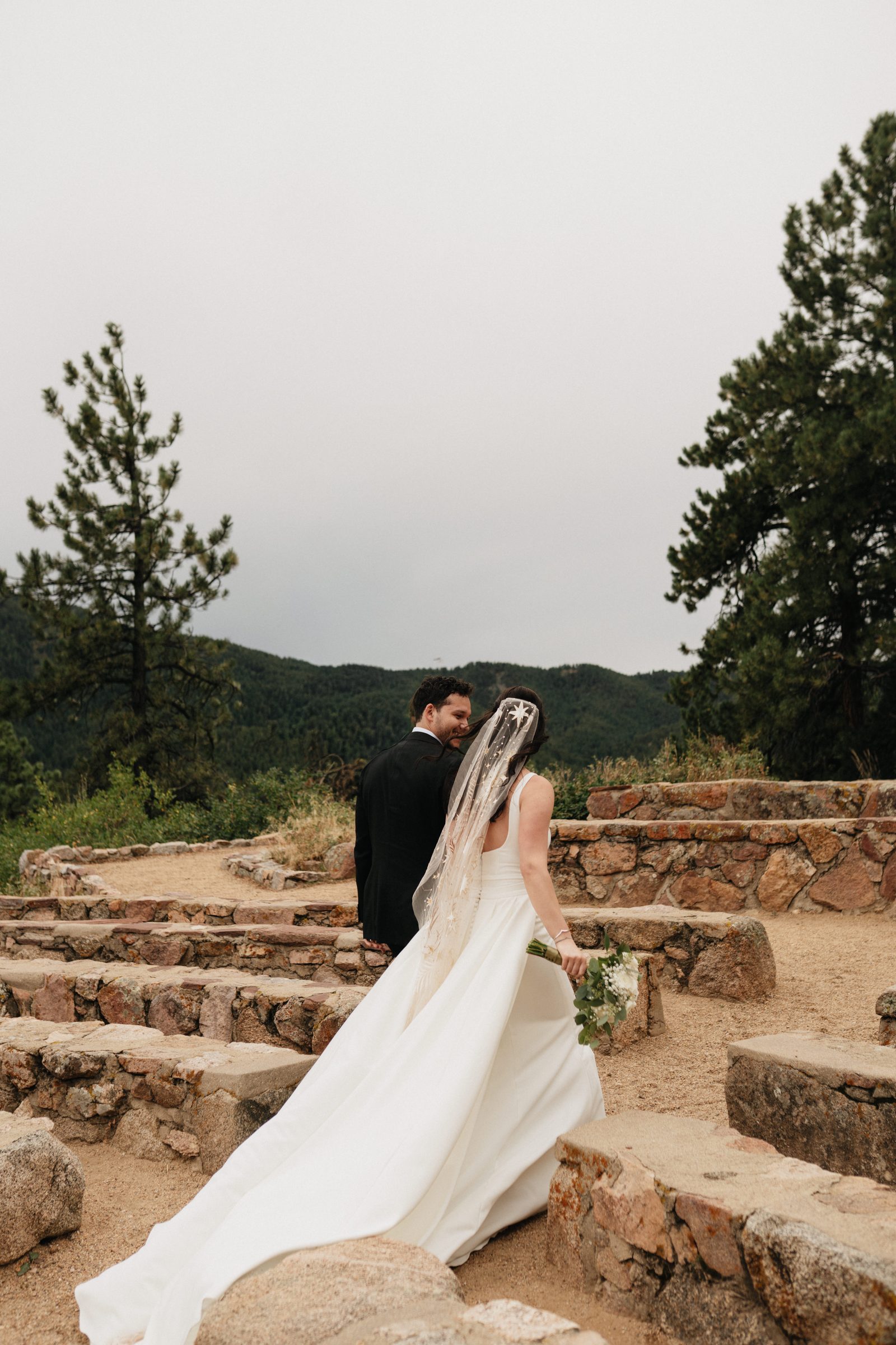 Bride in a long veil walks toward a stone amphitheater on a mountainside under a cloudy sky